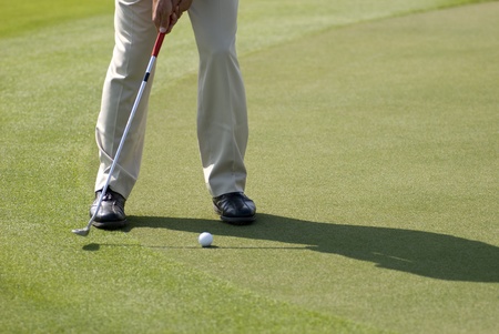 A golfer wearing beige pants and black leather shoes practicing his putt on a sunny day