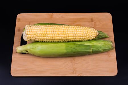 three unpeeled corn lie on a wooden board for cooking, background is black, top viewの写真素材
