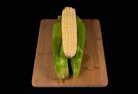 three unpeeled corn lie on a wooden board for cooking, background is black, top viewの写真素材