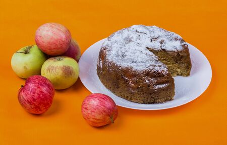 apple pie covered with icing sugar on a white plate, sliced cake, next to a bunch of ripe apples, photo on an orange background,の写真素材