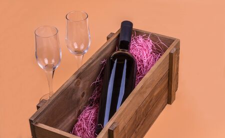 a bottle of red wine lies horizontally in a wooden box, next to it are two empty glasses for wine, background is beige, top view, close-upの写真素材