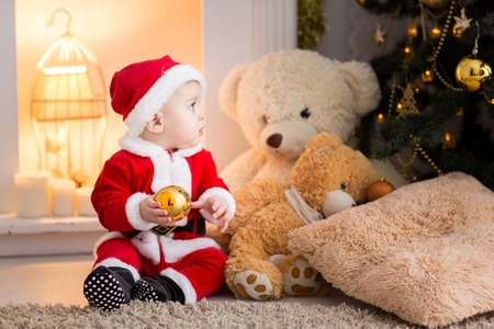 funny baby boy weared in Santa hat with golden Christmas ball, little Santa in the background of the Christmas treeの写真素材
