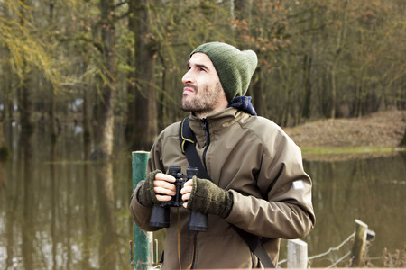 Man in jacket and binoculars standing on shore and observing riverの写真素材