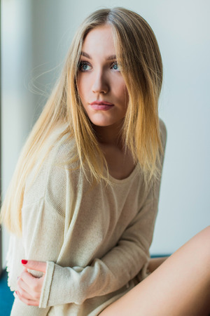Young blonde woman sitting next to window in sweater, looking emotive and thoughtfulの写真素材