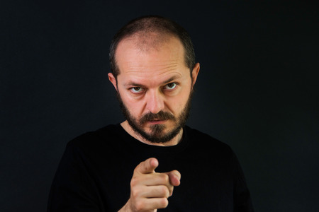Serious man with beard and mustaches on black background in low key, pointing finger towards cameraの写真素材