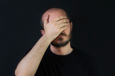 Serious man with beard and mustaches on black background, covering eyes with hand palm, face palm gestureの写真素材