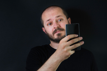 Man with beard and mustaches on black background in low key, holding smart phone and taking selfieの写真素材