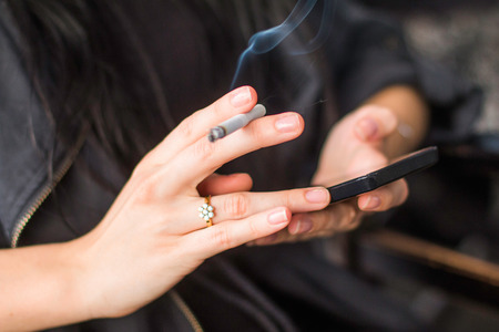 Young girl sitting in cafe, smoking cigarette and typing on smart phone, hands detailの写真素材