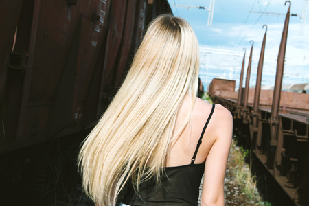 Beautiful blonde girl running between freight trains romantic look, from back