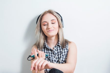Happy young girl in plaid shirt with headphones and smart phone sitting at floor, smiling and enjoying music at home. White wall, half body, youth music concept.の写真素材