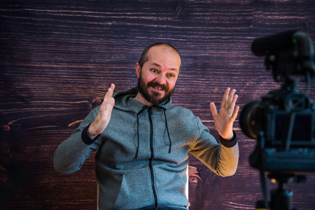 Bearded middle age man in hoody sitting and talking in front of DSLR camera, video blogging conceptの写真素材