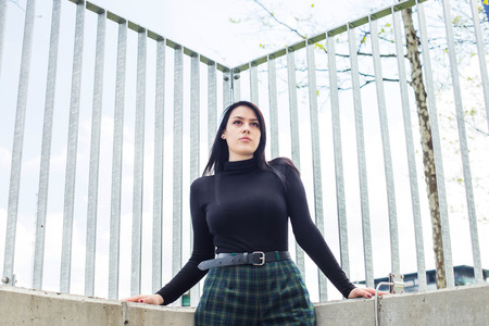 Young woman in black shirt and black hair standing next to fence outdoor in the city, half body, confidence concept, low perspectiveの写真素材