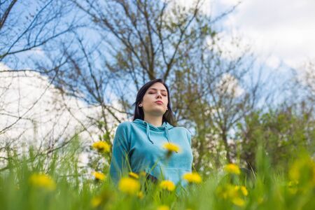Girl in hoodie sitting on green meadow among flowers and meditating in nature background. Woman practicing meditation. Eyes closed.の写真素材