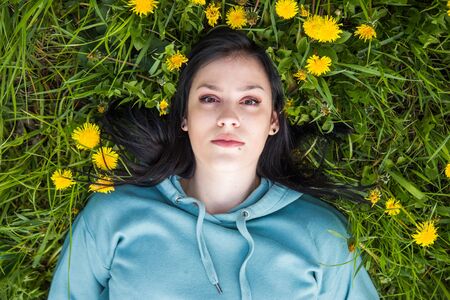 Head and shoulders portrait of beautiful young woman lying on the field in green grass and yellow dandelions, enjoying nature. Freedom and nature concept. Overhead shot.の写真素材