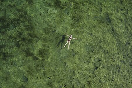 Woman in bikini floating on back on the surface of crystal clear water, young woman carefree on the sea, relaxing, Dugi otok island, Croatia, overhead drone viewの写真素材
