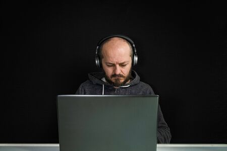 Caucasian man in hoodie and headphones sitting behind the laptop and typing, black backgroundの写真素材