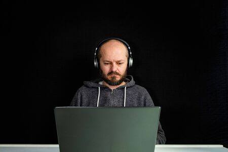 Caucasian man in hoodie and headphones sitting behind the laptop and typing, black backgroundの写真素材