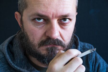 Adult bearded man in hoodie smoking cigarette, unhealthy habit, nicotine dependence closeup portrait, hand and lips detailの写真素材