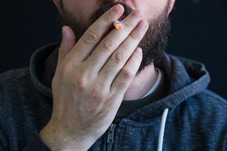 Adult bearded man in hoodie smoking cigarette, unhealthy habit, nicotine dependence closeup portrait, hand and lips detailの写真素材