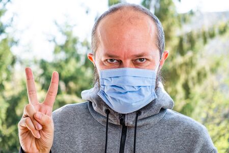 A bearded man with a mask shows a victory sign gesture with his fingers in nature park outdoorの写真素材