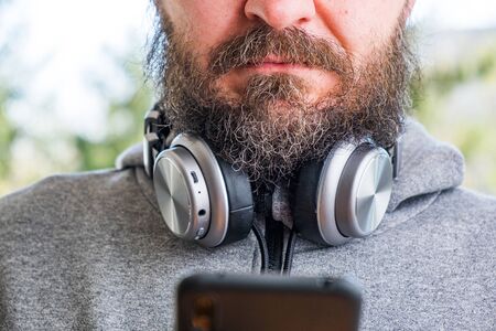 Bearded man in hoodie with wireless headphones around the neck using smart phone in park outdoor, reading or typing a message on smartphone, hands detailの写真素材