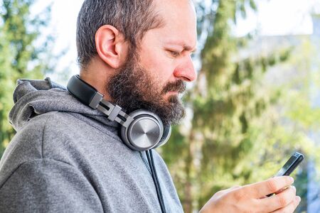 Bearded man in hoodie with wireless headphones around the neck using smart phone in park outdoor, reading or typing a message on smartphone, hands detailの写真素材