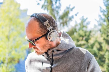Bearded man in hoodie and wireless headphones using smart phone in park outdoor, head and shouldersの写真素材