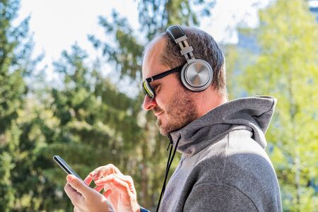 Bearded man in hoodie and wireless headphones using smart phone in park outdoor, head and shouldersの写真素材