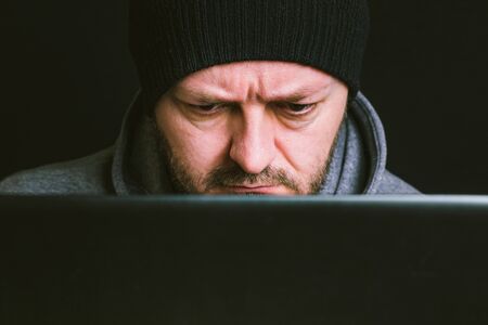 Man wearing black cap and hoodie sitting behind the computer monitor in the dark, looking at computer screen typing, close up, hacking computer system conceptの写真素材