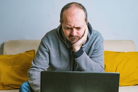 Young bearded man in casual clothes, trousers and hoodie sitting on a couch at the living room,...