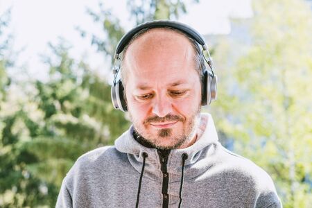 Bearded man in hoodie with headphones listening to music in park outdoors, head and shoulders