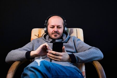 Bearded relaxed man in hoodie and trousers sitting on couch with headphones and listening music, smilingの写真素材