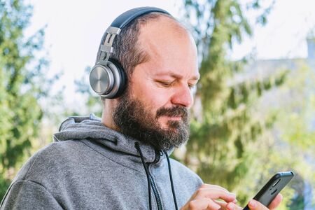 Bearded man in hoodie with headphones listening to music in park outdoors, head and shouldersの写真素材