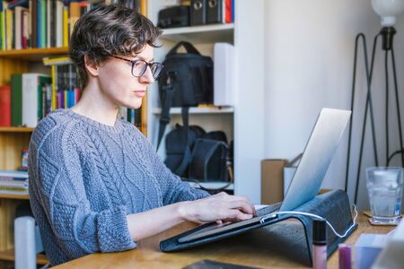 Woman working from home office. Young Caucasian short hair entrepreneur woman in sweater and glasses sitting and working online from home on computer laptop, candid, real photo, bookshelf behindの写真素材
