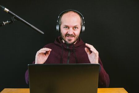 Guy in hoodie sitting behind the desk with laptop computer, headphones and microphone on black...
