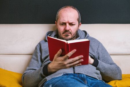 Serious young bearded man in casual clothes relaxed on couch reading red book on black backgroundの写真素材