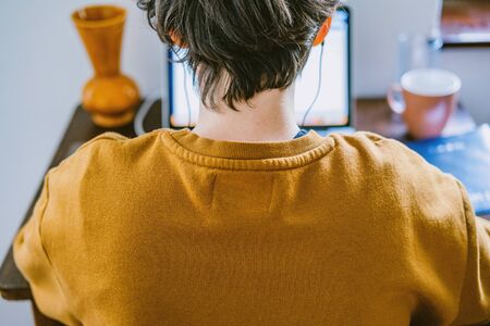 Woman working on desk in home office. Young Caucasian short hair woman with earphones working online from home on computer laptop from back, head and shoulders, real, candid photoの写真素材