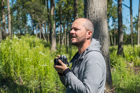 Adult man in hoodie walking in green forest with mirror less camera, beautiful summer day, half body portrait, landscape photography conceptの写真素材
