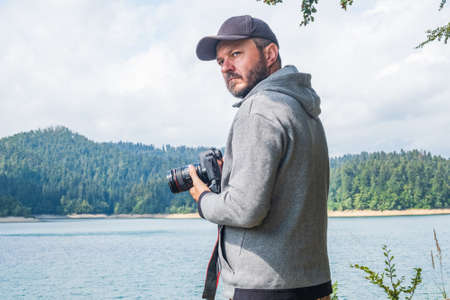 Adult man in hoodie walking around lake with DSLR camera ans shooting, beautiful summer day, half body portrait, landscape photography conceptの写真素材