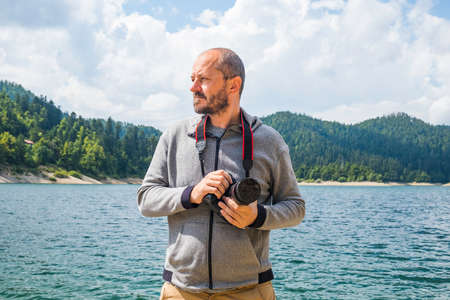 Photographer man in hoodie walking around lake with DSLR camera and shooting nature, half body portrait, landscape photography conceptの写真素材
