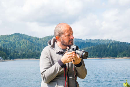 Photographer man in hoodie walking around lake with DSLR camera and shooting nature, half body portrait, landscape photography conceptの写真素材