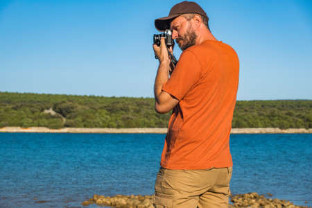 Photographer in t-shirt and cargo pants walking around sea shore with mirror less camera, shooting nature, half body portrait, landscape photography conceptの写真素材