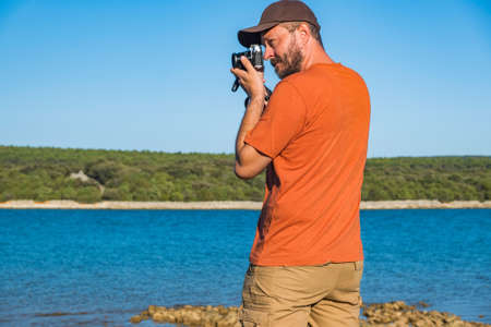 Photographer in t-shirt and cargo pants walking around sea shore with mirror less camera, shooting nature, half body portrait, landscape photography conceptの写真素材