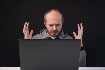 Man working from home office, angry face expression. Adult bearded sitting behind desk on black background, angry face, having business problem.の写真素材