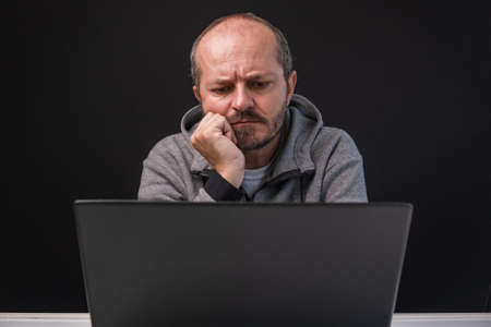 Worried man in hoodie working from home office. Adult bearded man sitting behind computer lap top, black background.の写真素材
