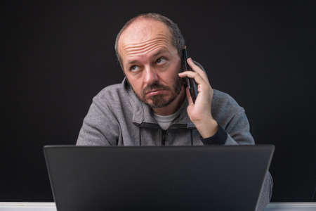 Bearded man in hoodie sitting behind lap top and talking on smart phoneの写真素材