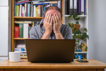 Man working online from home office on computer laptop behind vintage desk, with earphones, tired and exhausted after online meetingの写真素材