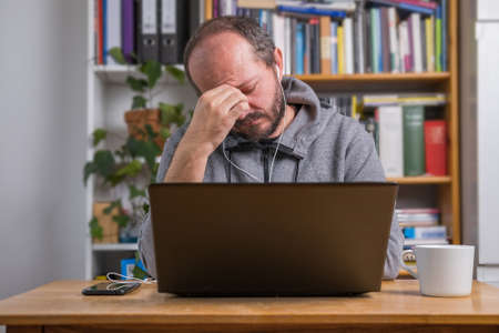 Man working online from home office on computer laptop behind vintage desk, with earphones, tired and exhausted after online meeting, facepalm expressionの写真素材