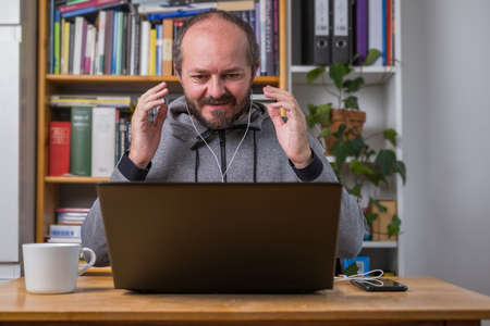 Bearded man working online from home on computer laptop behind vintage desk, with earphones, talking and explaining to colleagues on online meetingの写真素材