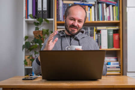 Bearded man working online from home on computer laptop behind vintage desk, with earphones, talking and explaining to colleagues on online meetingの写真素材